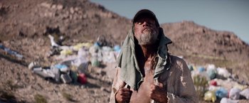 Movie still from “The Bad Batch” (2016), directed by Ana Lily Amirpour – An old man is standing in the dirt with his hands on his chest; Medium shot, Low angle