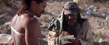 Movie still from “The Bad Batch” (2016), directed by Ana Lily Amirpour – A man writing on a notebook while another man looks on; Medium shot, Over the shoulder angle