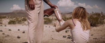 Movie still from “The Bad Batch” (2016), directed by Ana Lily Amirpour – A person with a broken arm is being helped by another person; Medium shot, Over the shoulder angle