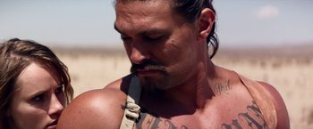 Movie still from “The Bad Batch” (2016), directed by Ana Lily Amirpour – A man with a knife in his mouth on the beach; Close Up shot, Over the shoulder angle