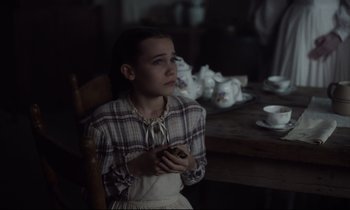 Movie still from “The Beguiled” (2017), directed by Sofia Coppola – A girl sitting in a chair in front of a wooden table; Medium shot, High angle