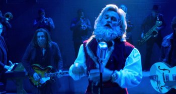 Movie still from “The Christmas Chronicles” (2018), directed by Clay Kaytis – A man dressed as santa claus singing into a microphone in front of a group of people; Medium shot, Over the shoulder angle