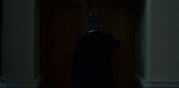 Movie still from “The Crown” (2016), created by Peter Morgan – A man standing in front of a door in a dark room; Medium shot, Low angle