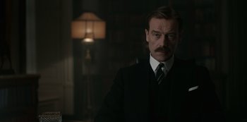 Movie still from “The Crown” (2016), created by Peter Morgan – A man in a suit and tie standing in front of a lamp; Close Up shot, Low angle