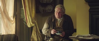 Movie still from “Mr. Turner” (2014), directed by Mike Leigh – An older man sitting in a chair holding a bowl; Medium shot, Over the shoulder angle