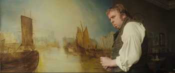 Movie still from “Mr. Turner” (2014), directed by Mike Leigh – A man standing in front of a painting of boats in the water; Medium shot, Over the shoulder angle