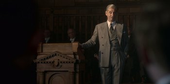 Movie still from “The Crown” (2016), created by Peter Morgan – An older man in a suit is standing at a lectern; Medium shot, Low angle