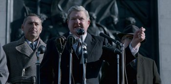 Movie still from “The Crown” (2016), created by Peter Morgan – A man in a suit is speaking into a microphone; Medium shot, Over the shoulder angle