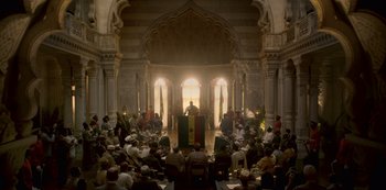 Movie still from “The Crown” (2016), created by Peter Morgan – A crowd of people sitting in a large room; Extreme Wide shot, High angle