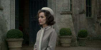 Movie still from “The Crown” (2016), created by Peter Morgan – A woman wearing a white hat standing in front of a building; Medium shot, Over the shoulder angle