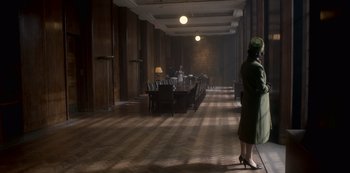 Movie still from “The Crown” (2016), created by Peter Morgan – A woman in a green coat standing in front of a long table; Wide shot, High angle