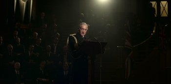 Movie still from “The Crown” (2016), created by Peter Morgan – A man in a military uniform is speaking at a podium; Medium shot, Low angle