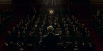 Movie still from “The Crown” (2016), created by Peter Morgan – A group of men sitting in front of an audience; Wide shot, High angle