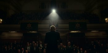 Movie still from “The Crown” (2016), created by Peter Morgan – A man is standing in front of a crowd of people; Wide shot, Low angle