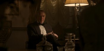 Movie still from “The Crown” (2016), created by Peter Morgan – An older man in a tuxedo sitting in front of a lamp; Medium shot, Over the shoulder angle