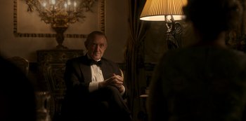Movie still from “The Crown” (2016), created by Peter Morgan – An older man in a tuxedo sitting in front of a lamp; Medium shot, Over the shoulder angle