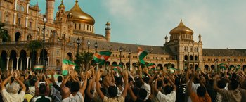 Movie still from “The Dictator” (2012), directed by Larry Charles – A group of people standing in front of a building with their hands up; Extreme Wide shot, High angle