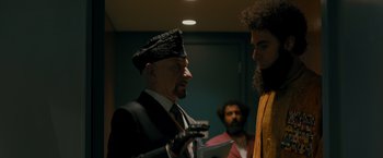 Movie still from “The Dictator” (2012), directed by Larry Charles – An older man wearing a hat and holding a microphone; Medium shot, Over the shoulder angle