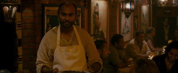 Movie still from “The Dictator” (2012), directed by Larry Charles – A man in an apron is holding a tray of food; Medium shot, Over the shoulder angle
