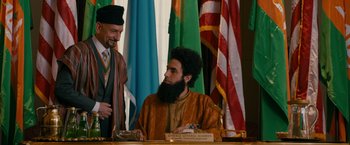 Movie still from “The Dictator” (2012), directed by Larry Charles – A man with a long beard sitting at a table with other people; Medium shot, Over the shoulder angle