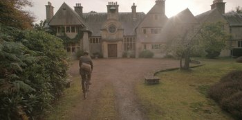 Movie still from “The Dig” (2021), directed by Simon Stone – A man riding a bike down a dirt road near a house; Extreme Wide shot, Low angle