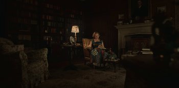 Movie still from “The Dig” (2021), directed by Simon Stone – A woman sitting in a chair reading a book in a dark room; Wide shot, High angle