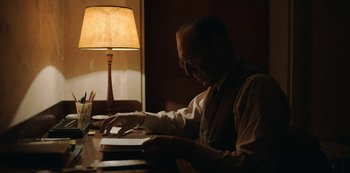 Movie still from “The Dig” (2021), directed by Simon Stone – An older man sitting at a table with a book; Medium shot, Over the shoulder angle
