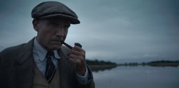 Movie still from “The Dig” (2021), directed by Simon Stone – An older man smoking a pipe while standing in front of a body of water; Close Up shot, Low angle