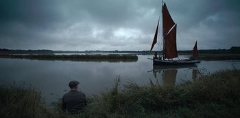 Movie still from “The Dig” (2021), directed by Simon Stone – A man sitting on the shore of a body of water; Wide shot, High angle