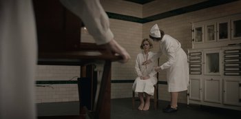 Movie still from “The Dig” (2021), directed by Simon Stone – A woman sitting on a chair in front of a nurse; Medium shot, High angle