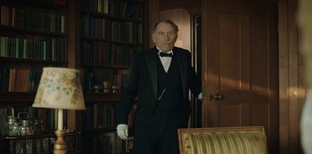 Movie still from “The Dig” (2021), directed by Simon Stone – An older man in a tuxedo standing in front of bookshelves; Medium shot, Low angle