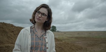 Movie still from “The Dig” (2021), directed by Simon Stone – A woman wearing glasses and a plaid shirt standing in the middle of an open field; Medium shot, Over the shoulder angle