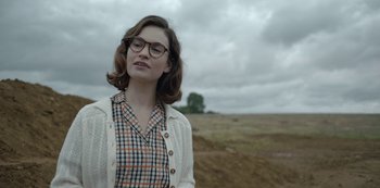 Movie still from “The Dig” (2021), directed by Simon Stone – A woman wearing glasses and a plaid shirt standing in the middle of an open field; Medium shot, Over the shoulder angle