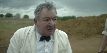 Movie still from “The Dig” (2021), directed by Simon Stone – An older man in a white jacket and a black bow tie; Close Up shot, Over the shoulder angle