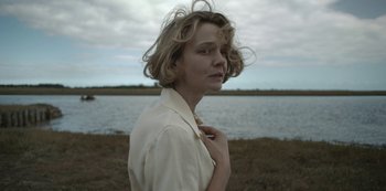 Movie still from “The Dig” (2021), directed by Simon Stone – A woman in a white shirt standing next to a body of water; Close Up shot, High angle