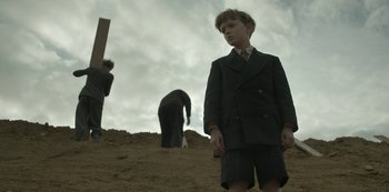 Movie still from “The Dig” (2021), directed by Simon Stone – A young boy in a suit stands in the sand; Medium shot, Low angle