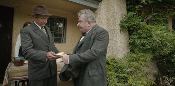 Movie still from “The Dig” (2021), directed by Simon Stone – A man in a suit and hat is holding a sandwich; Medium shot, Over the shoulder angle