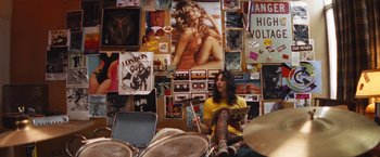 Movie still from “The Dirt” (2019), directed by Jeff Tremaine – A woman sitting on a drum set in front of a wall covered with photos; Medium shot, Overhead angle