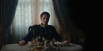 Movie still from “The Empress” (2022), created by Katharina Eyssen – A man sitting at a table with a plate of food on it; Medium shot, Low angle