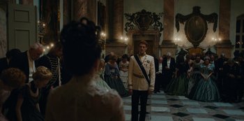 Movie still from “The Empress” (2022), created by Katharina Eyssen – A man in a white suit stands in front of a group of people; Wide shot, Over the shoulder angle