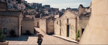 Movie still from “No Time to Die” (2021), directed by Cary Joji Fukunaga – A person riding a motorcycle down a street near some buildings; Extreme Wide shot, High angle