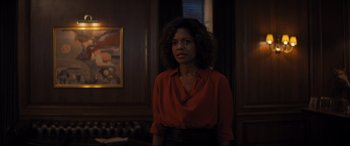 Movie still from “No Time to Die” (2021), directed by Cary Joji Fukunaga – A woman in a red shirt is standing in front of a painting; Medium shot, Low angle