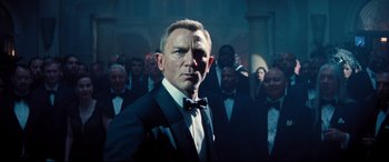 Movie still from “No Time to Die” (2021), directed by Cary Joji Fukunaga – A man in a suit and bow tie standing in front of a group of men; Medium shot, Low angle