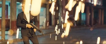 Movie still from “No Time to Die” (2021), directed by Cary Joji Fukunaga – A man in a black suit holding a gun in the street; Medium shot, Low angle