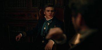 Movie still from “The Empress” (2022), created by Katharina Eyssen – A young man sitting in a chair wearing a suit and tie; Medium shot, Over the shoulder angle