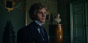 Movie still from “The Empress” (2022), created by Katharina Eyssen – A man in a suit and tie standing in front of a statue; Close Up shot, Low angle