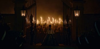 Movie still from “The Empress” (2022), created by Katharina Eyssen – A group of people standing around a fire pit at night; Extreme Wide shot, Over the shoulder angle