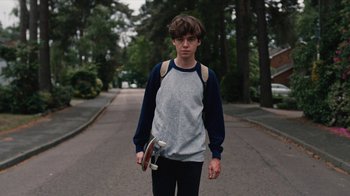 Movie still from “The End of the F***ing World” (2017), directed by Jonathan Entwistle – A young man holding a skateboard while standing on the side of the road; Medium shot, Low angle