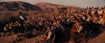 Movie still from “Babylon” (2022), directed by Damien Chazelle – A large group of people sitting in a field with musical instruments; Extreme Wide shot, High angle