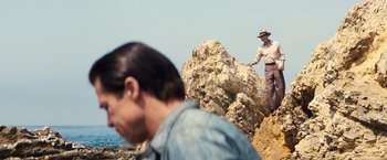 Movie still from “Babylon” (2022), directed by Damien Chazelle – A man standing on top of a rock near a body of water; Wide shot, Over the shoulder angle